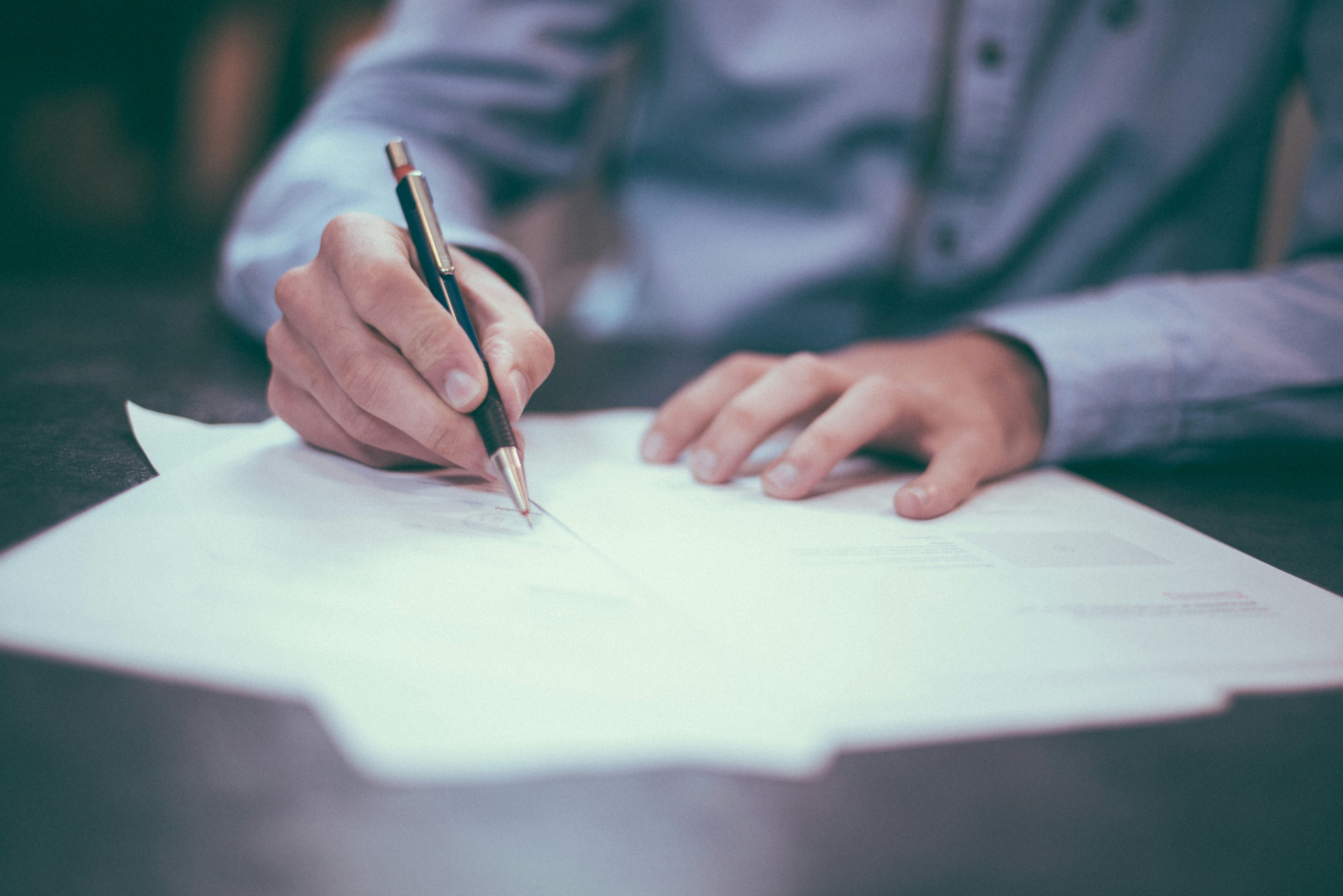 Image of hands; one of them is holidng a pen and writing on a piece of paper whilst the other is resting on a desk. This symbolises the process of drafting a policy.
